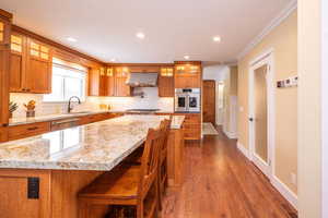Kitchen featuring brown cabinetry, glass insert cabinets, recessed lighting, crown molding, and light stone counters