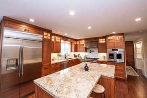 Kitchen featuring brown cabinetry, appliances with stainless steel finishes, light stone countertops, glass insert cabinets, and ornamental molding