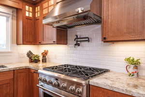 Kitchen featuring brown cabinets, ventilation hood, stainless steel gas stove, backsplash, and light stone countertops