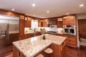 Kitchen with brown cabinetry, appliances with stainless steel finishes, glass insert cabinets, light stone counters, and recessed lighting