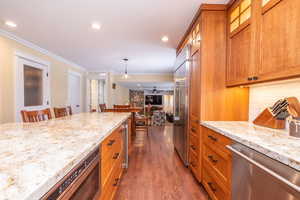 Kitchen featuring brown cabinets, ornamental molding, stainless steel appliances, light stone counters, and dark wood finished floors