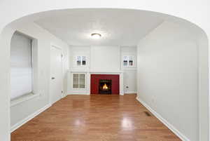 Unfurnished living room featuring arched walkways, a textured ceiling, wood finished floors, and a brick fireplaceFireplace flame image  is virtual
