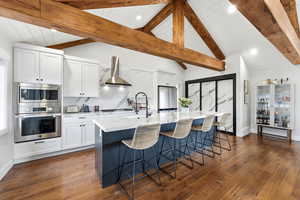 Kitchen with white cabinets, appliances with stainless steel finishes, lofted ceiling, an island with sink, and wall chimney exhaust hood