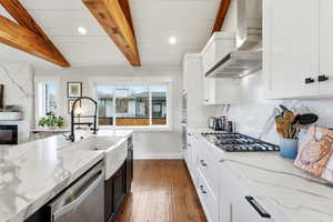 Kitchen with light stone countertops, white cabinets, appliances with stainless steel finishes, wall chimney exhaust hood, and beam ceiling