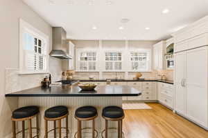 Kitchen with dark countertops, a peninsula, paneled built in refrigerator, a kitchen breakfast bar, and wall chimney range hood