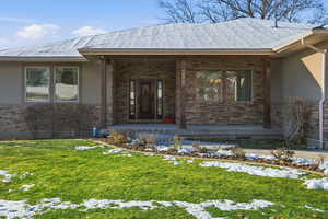 Snow covered property entrance with covered porch, a lawn, stucco siding, stone siding, and roof with shingles