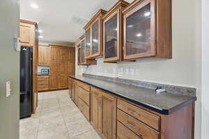 Kitchen with brown cabinetry, dark countertops, freestanding refrigerator, light tile patterned flooring, and recessed lighting