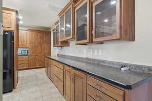 Kitchen with brown cabinets, freestanding refrigerator, dark countertops, light tile patterned flooring, and glass insert cabinets