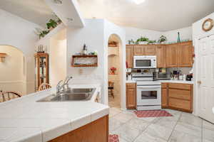 Kitchen with arched walkways, tile counters, white appliances, lofted ceiling, and brown cabinetry