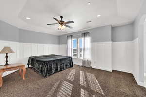 Carpeted bedroom featuring ceiling fan, a wainscoted wall, a tray ceiling, a decorative wall, and recessed lighting