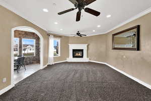 Unfurnished living room with ornamental molding, arched walkways, a glass covered fireplace, carpet flooring, and a ceiling fan