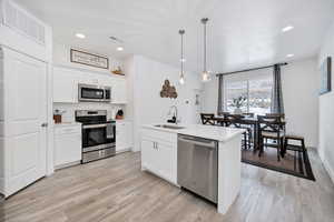 Kitchen featuring appliances with stainless steel finishes, decorative light fixtures, white cabinets, an island with sink, and light wood-type flooring