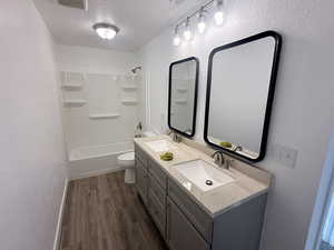 Bathroom featuring a textured wall, double vanity, bathing tub / shower combination, a textured ceiling, and dark wood finished floors
