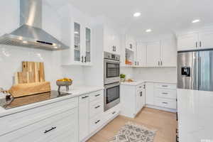 Kitchen featuring white cabinets, wall chimney range hood, appliances with stainless steel finishes, light stone countertops, and light wood-type flooring