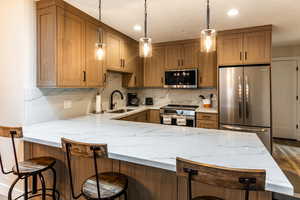Kitchen featuring light stone countertops, stainless steel appliances, hanging light fixtures, brown cabinetry, and recessed lighting