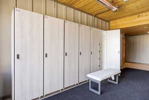 Locker room featuring multiple light wood-finish lockers with individual locks, a padded bench, and a textured dark gray floor covering