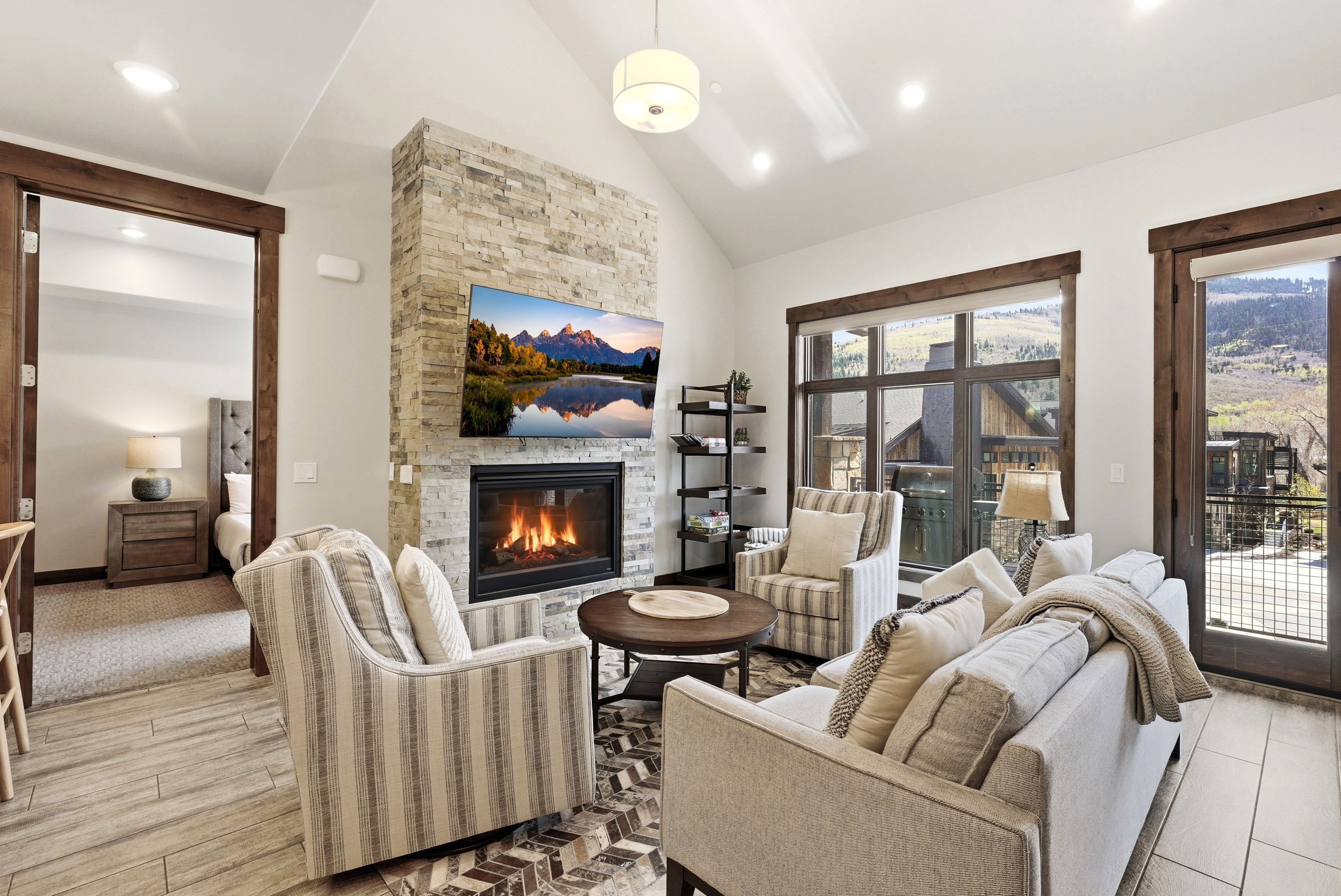 Living area featuring a stone-clad fireplace with a mounted television, a vaulted ceiling, wood-finish flooring, a wall of windows, and a glass-paned door with dark wood trim