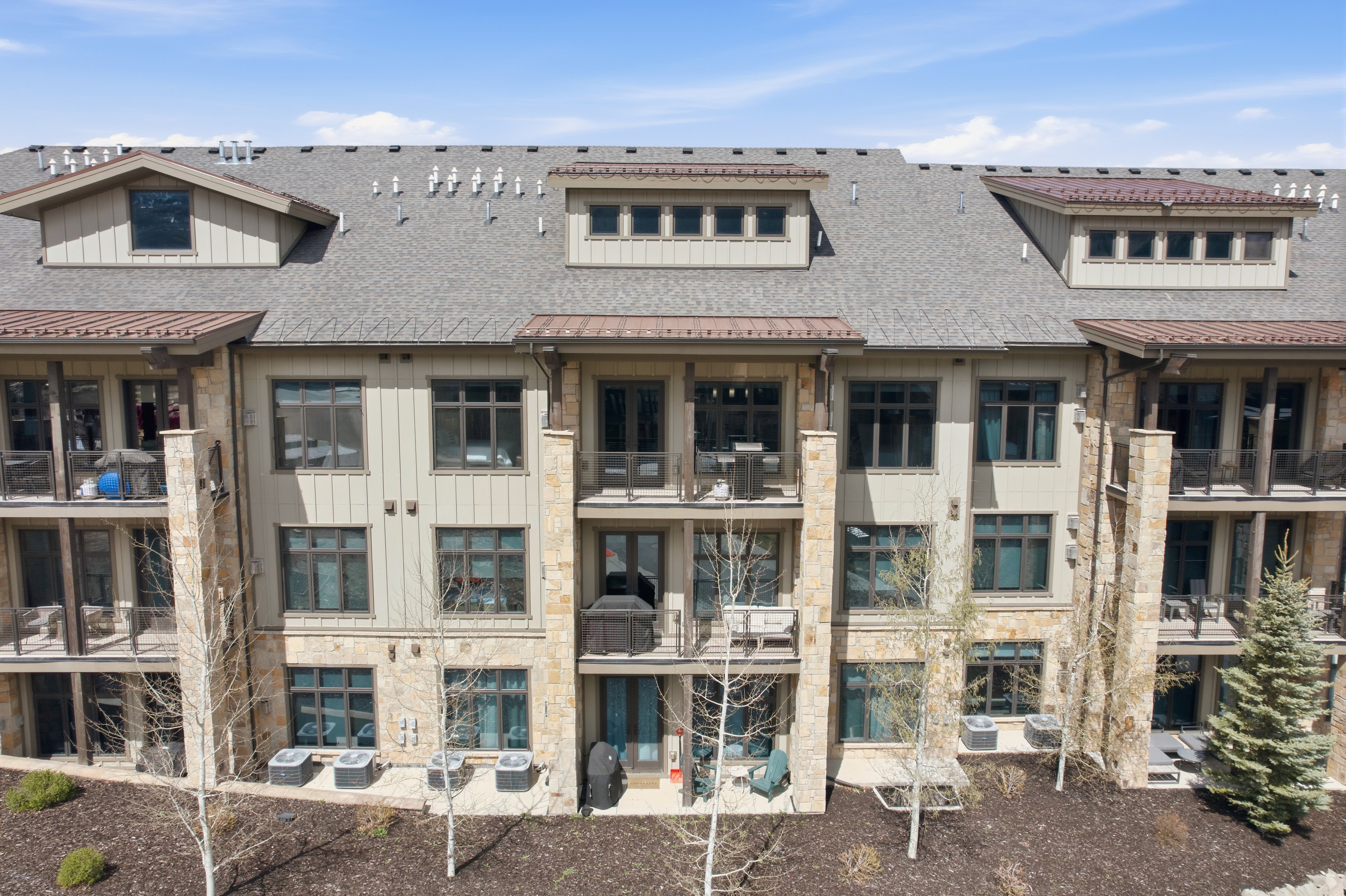 Multi-story condominium building featuring stone and siding exterior, multiple balconies, individual patios, and dormer windows