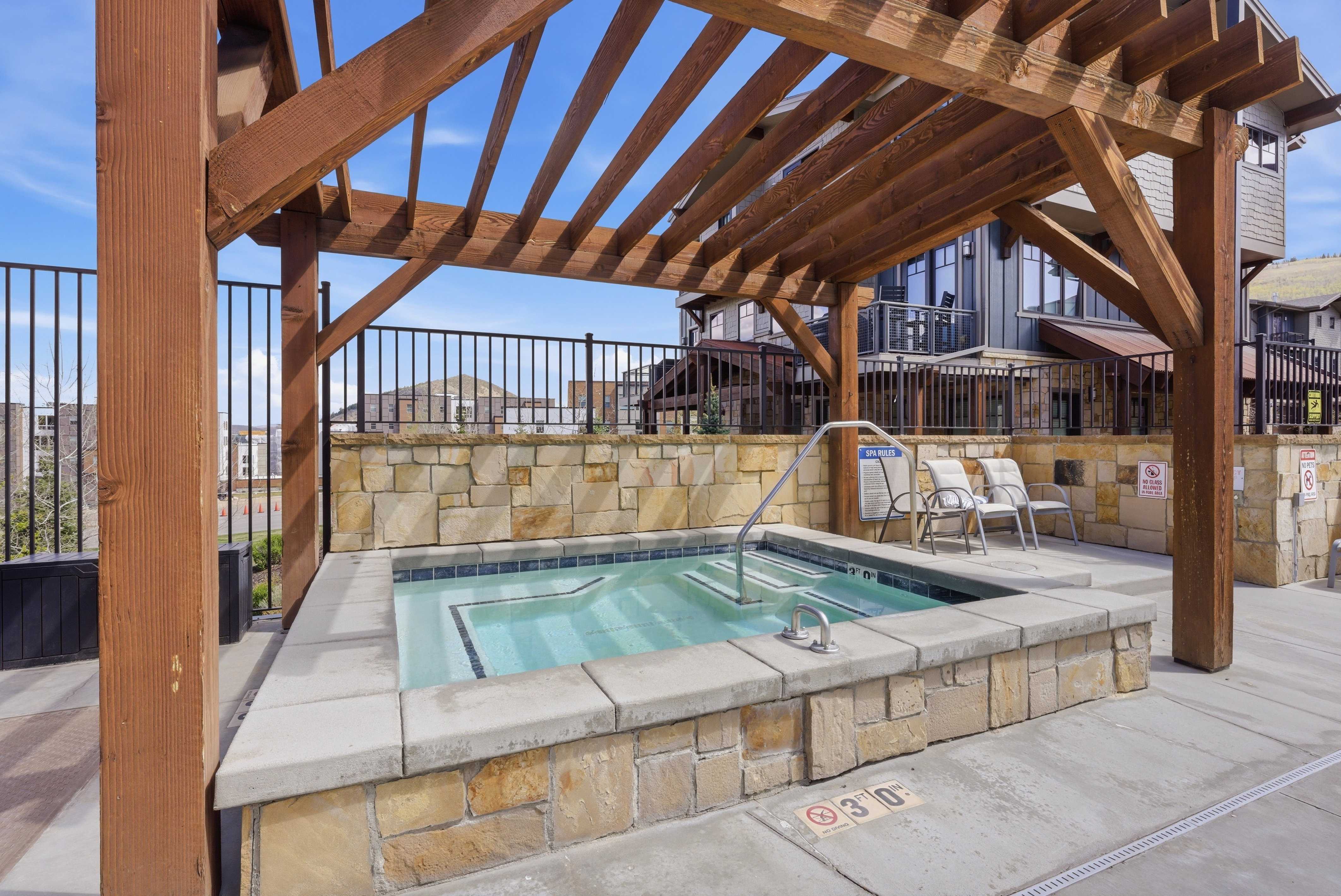 Outdoor spa with stone surround, concrete patio, wood pergola, and black metal fencing