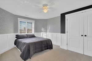 Carpeted bedroom featuring wallpapered walls, wainscoting, a closet, ceiling fan, and a decorative wall