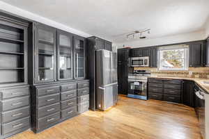 Kitchen featuring appliances with stainless steel finishes, glass insert cabinets, light wood-type flooring, a textured ceiling, and open shelves