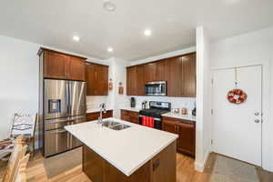 Kitchen with stainless steel appliances, an island with sink, recessed lighting, light wood-type flooring, and light stone countertops