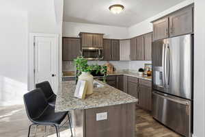 Kitchen with appliances with stainless steel finishes, light stone counters, dark wood-type flooring, dark brown cabinetry, and a kitchen island with sink