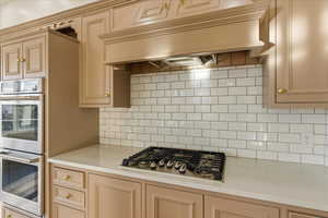 Kitchen with premium range hood, stainless steel appliances, light stone countertops, cream cabinets, and tasteful backsplash