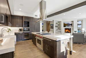 Kitchen featuring dark brown cabinetry, island range hood, modern cabinets, light stone countertops, and beamed ceiling
