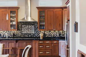 Kitchen featuring wall chimney exhaust hood, freestanding refrigerator, brown cabinets, and glass insert cabinets