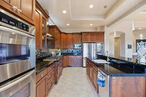 Kitchen featuring appliances with stainless steel finishes, dark stone counters, brown cabinetry, recessed lighting, and glass insert cabinets