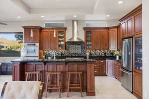 Kitchen with a breakfast bar, an island with sink, stainless steel appliances, wall chimney exhaust hood, and brown cabinets