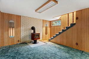 Unfurnished living room with stairway, carpet flooring, a wood stove, wooden walls, and a textured ceiling