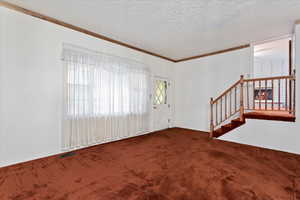 Unfurnished living room featuring a textured ceiling, dark colored carpet, stairway, and crown molding