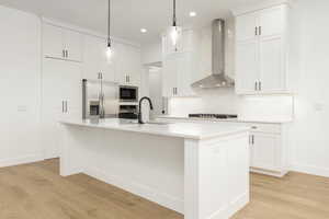 Kitchen featuring wall chimney range hood, stainless steel appliances, backsplash, a center island with sink, and light wood-style floors