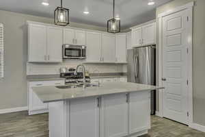 Kitchen featuring white cabinetry, a center island with sink, stainless steel appliances, light stone countertops, and decorative light fixtures