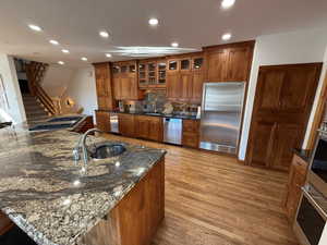 Kitchen with brown cabinets, glass insert cabinets, stainless steel appliances, dark stone countertops, and light wood finished floors