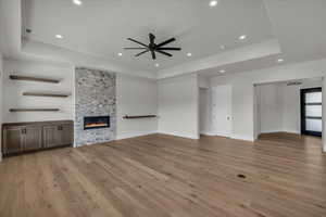 Unfurnished living room with a tray ceiling, a stone fireplace, light wood-style flooring, ceiling fan, and recessed lighting