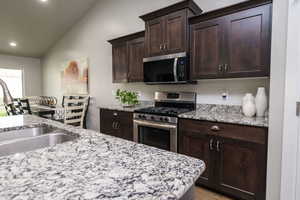 Kitchen with dark brown cabinetry, appliances with stainless steel finishes, light stone countertops, lofted ceiling, and recessed lighting