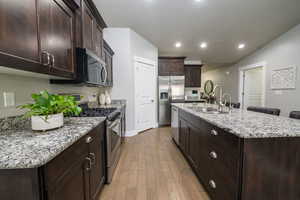 Kitchen with stainless steel appliances, dark brown cabinets, a center island with sink, light stone counters, and light wood-type flooring