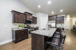 Kitchen with appliances with stainless steel finishes, dark brown cabinetry, light stone countertops, an island with sink, and a breakfast bar area