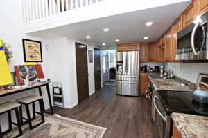 Kitchen with appliances with stainless steel finishes, dark wood-type flooring, recessed lighting, brown cabinetry, and light stone countertops