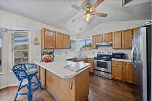 Kitchen featuring a peninsula, appliances with stainless steel finishes, brown cabinets, vaulted ceiling, and dark wood finished floors