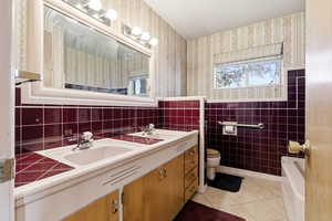 Full bathroom featuring wallpapered walls, double vanity, light tile patterned floors, tile walls, and wainscoting
