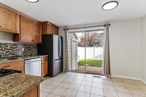 Kitchen featuring dark stone counters, brown cabinetry, stainless steel appliances, tasteful backsplash, and light tile patterned floors