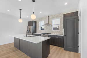 Kitchen featuring decorative light fixtures, a kitchen island with sink, wall chimney range hood, stainless steel appliances, and tasteful backsplash
