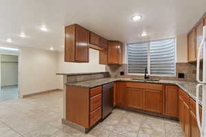 Kitchen featuring brown cabinetry, dark stone counters, decorative backsplash, dishwasher, and recessed lighting