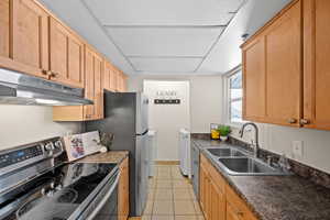 Kitchen featuring appliances with stainless steel finishes, under cabinet range hood, dark countertops, washing machine and clothes dryer, and light tile patterned floors
