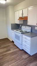 Kitchen featuring white electric range, dark wood finished floors, white cabinets, under cabinet range hood, and dark countertops