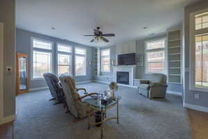 Carpeted living room with a glass covered fireplace, built in shelves, and a ceiling fan